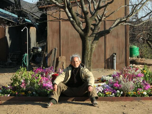 険しい表情で座り込む夏八木勲扮する小野泰彦